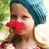 A young child with blonde hair, wearing a Molly Beret from Never Not Knitting in blue and white stripes with a lace brim, holds and smells a red flower. The child is dressed in a colorful floral-patterned dress. The background shows greenery and is brightly lit.