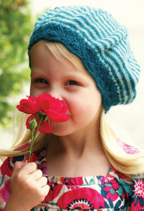 A young child with blonde hair, wearing a Molly Beret from Never Not Knitting in blue and white stripes with a lace brim, holds and smells a red flower. The child is dressed in a colorful floral-patterned dress. The background shows greenery and is brightly lit.