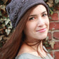 A woman with long brown hair is wearing a Plum Tree Slouch Hat by Never Not Knitting, featuring a charming knitted leaf pattern. She pairs it with a grey top and stands in front of an ivy-covered brick wall, smiling softly and looking towards the camera.