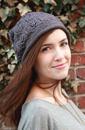 A woman with long brown hair is wearing a Plum Tree Slouch Hat by Never Not Knitting, featuring a charming knitted leaf pattern. She pairs it with a grey top and stands in front of an ivy-covered brick wall, smiling softly and looking towards the camera.