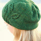 A person with long, blonde hair is wearing the Seedling Beanie from Never Not Knitting. The green knitted hat resembles a slouchy beanie and features a pattern of leaves on one side, reminiscent of fledgling plants. The background is blurred to emphasize the beanie and the person's hair.