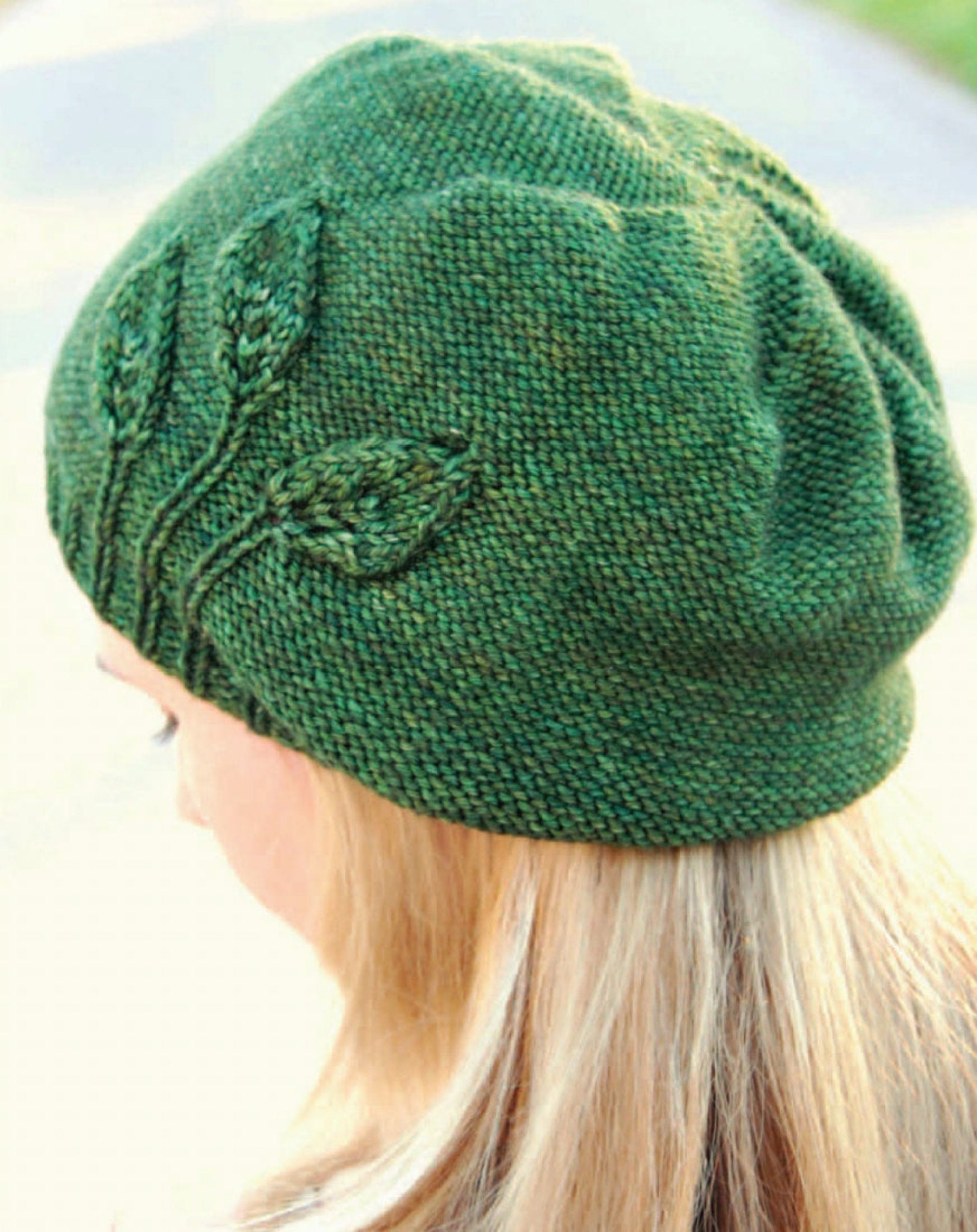 A person with long, blonde hair is wearing the Seedling Beanie from Never Not Knitting. The green knitted hat resembles a slouchy beanie and features a pattern of leaves on one side, reminiscent of fledgling plants. The background is blurred to emphasize the beanie and the person's hair.