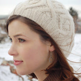 A woman with light skin and brown hair is wearing the Winter Trails Hat by Never Not Knitting, a beige knitted hat that’s the perfect accessory for a cold winter's day. She also has dangling earrings and another warm cabled hat. She is looking to the left against a backdrop of a snowy outdoor setting, while donning a cozy brown sweater.