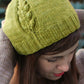 A person is pictured outdoors, wearing the Perennial Hat by Never Not Knitting. The hat features beautiful cable knit details in a green hue. The person, with long brown hair and dressed in a cozy brown sweater, is looking down while adjusting the beanie with both hands. Blurred greenery in the background adds a serene touch to the scene.