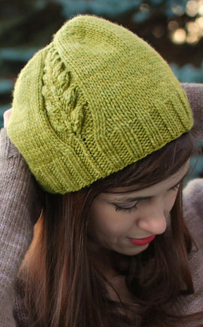 A person is pictured outdoors, wearing the Perennial Hat by Never Not Knitting. The hat features beautiful cable knit details in a green hue. The person, with long brown hair and dressed in a cozy brown sweater, is looking down while adjusting the beanie with both hands. Blurred greenery in the background adds a serene touch to the scene.