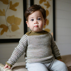 A young child with brown hair sits on a beige surface, wearing the Tot Toppers Baby Tee from Never Not Knitting, featuring beige and olive stripes made from fingering weight yarn, paired with light grey pants. Behind the child, two framed botanical prints with illustrations of leaves are mounted on a white wall.