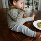 A young child with brown hair, wearing a grey and brown striped Tot Toppers Baby Tee by Never Not Knitting and grey pants, sits on a wooden table. The child is holding a cookie and eating it, with a white plate of cookies and a glass of milk nearby, looking cozy as if in a knitted baby onesie.