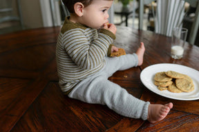 A young child with brown hair, wearing a grey and brown striped Tot Toppers Baby Tee by Never Not Knitting and grey pants, sits on a wooden table. The child is holding a cookie and eating it, with a white plate of cookies and a glass of milk nearby, looking cozy as if in a knitted baby onesie.