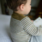 A young child with short brown hair sits on a bed while holding and looking at a book. The child is wearing the Tot Toppers Baby Tee by Never Not Knitting, made from fingering weight yarn, paired with grey pants. They are facing away from the camera, with their head slightly turned to the right.