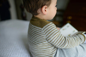 A young child with short brown hair sits on a bed while holding and looking at a book. The child is wearing the Tot Toppers Baby Tee by Never Not Knitting, made from fingering weight yarn, paired with grey pants. They are facing away from the camera, with their head slightly turned to the right.