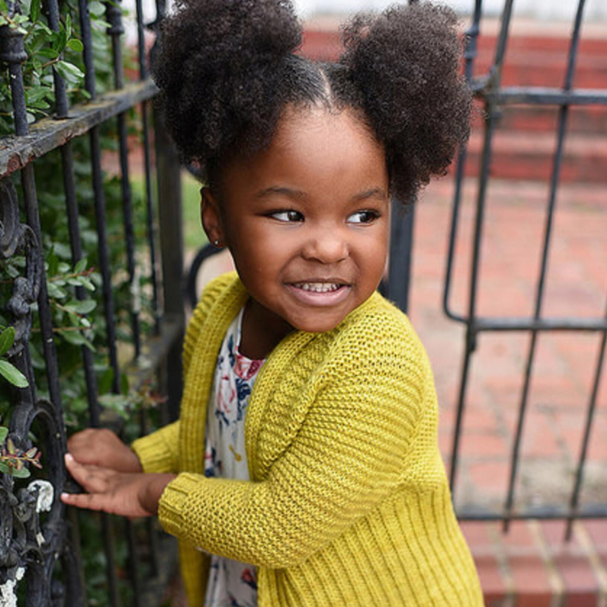 A young girl, adorned in the Tot Toppers Little Sister cardigan downloaded from Never Not Knitting, smiles brightly near a wrought-iron gate. She has two puffy ponytails and is looking to the side. The background features lush green leaves and classic red bricks.