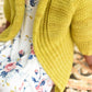 A person wearing a Tot Toppers Little Sister cardigan by Never Not Knitting, crafted with DK weight yarn in yellow, sits over a white dress adorned with a colorful floral pattern on an ornate, vintage-style metal bench. The focus is on the outfit, emphasizing the contrasting textures and colors.