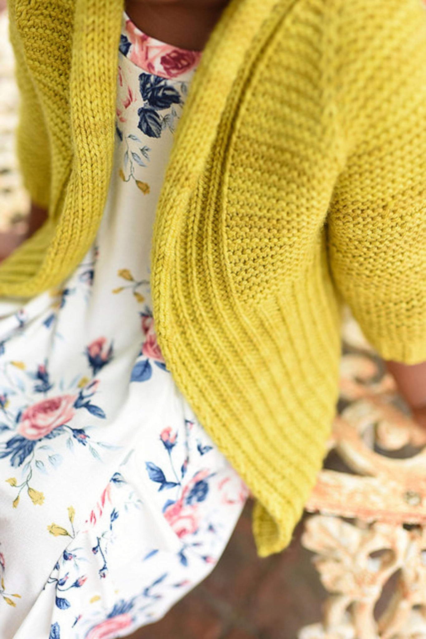 A person wearing a Tot Toppers Little Sister cardigan by Never Not Knitting, crafted with DK weight yarn in yellow, sits over a white dress adorned with a colorful floral pattern on an ornate, vintage-style metal bench. The focus is on the outfit, emphasizing the contrasting textures and colors.