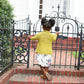 A young child wearing a colorful Tot Toppers Little Sister cardigan by Never Not Knitting, paired with a floral dress, stands in front of an ornate black iron gate nestled within a hedge. The kid's yellow sweater, meticulously crafted with the elegance of DK weight yarn, accentuates their charm as they look up at the closed gate with hands gently placed on it. In the background, a white building with a door can be seen.