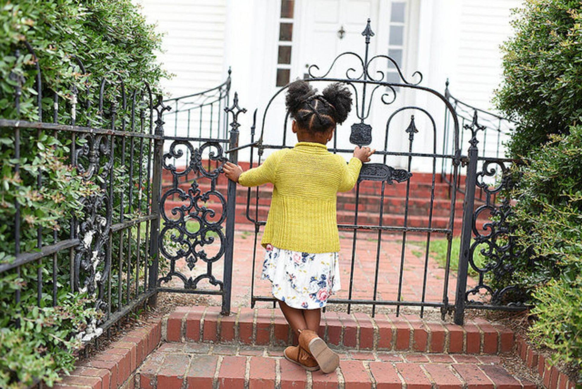 A young child with her hair in pigtails is wearing the Tot Toppers Little Sister cardigan from Never Not Knitting, along with a white dress featuring a floral pattern. She is standing at an ornate black wrought-iron gate, which leads to a house with white siding and a red-brick pathway. Bushes line both sides of the gate.