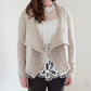 A person stands against a plain white background, wearing the Knitbot Harborside Cardigan by Never Not Knitting. The advanced beginner's worsted weight cardigan features a wide, draped collar and is worn over a white lace top and dark pants. Their hands are relaxed by their sides, and their head is cropped out of the frame.