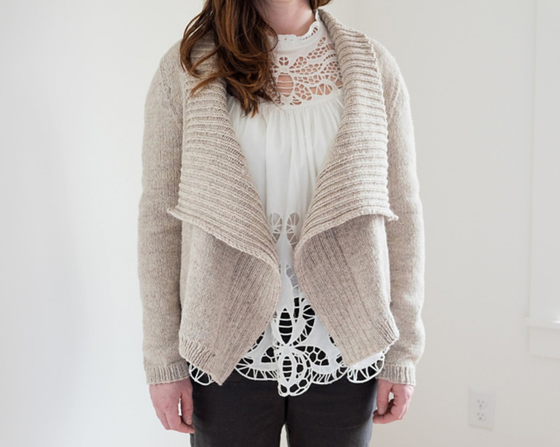 A person stands against a plain white background, wearing the Knitbot Harborside Cardigan by Never Not Knitting. The advanced beginner's worsted weight cardigan features a wide, draped collar and is worn over a white lace top and dark pants. Their hands are relaxed by their sides, and their head is cropped out of the frame.