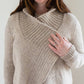 A person wearing the Knitbot Harborside Cardigan from Never Not Knitting, a beige textured sweater with a distinctive overlapping collar, holds the edge of the collar with one hand. The cardigan, reminiscent of an advanced beginner's knitting pattern, showcases a mix of ribbed and smooth knit patterns. The person's other hand, also visible, is adorned with a ring.