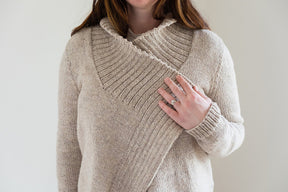 A person wearing the Knitbot Harborside Cardigan from Never Not Knitting, a beige textured sweater with a distinctive overlapping collar, holds the edge of the collar with one hand. The cardigan, reminiscent of an advanced beginner's knitting pattern, showcases a mix of ribbed and smooth knit patterns. The person's other hand, also visible, is adorned with a ring.