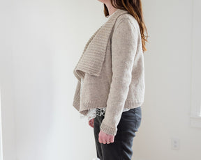 A person with long brown hair is standing indoors, facing away from the camera. They are wearing the light beige Knitbot Harborside Cardigan by Never Not Knitting, featuring a worsted weight knit and a waterfall front, layered over a white patterned top and paired with dark pants. The background is plain with a light-colored wall and an electrical outlet visible. Perfect inspiration for advanced beginner knitters!