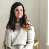 A woman with long, dark hair and a hint of a smile sits on a wooden chair, wearing the Knitbot Harborside Cardigan by Never Not Knitting over a white lace-detailed top. The cardigan is made using textured, off-white worsted weight yarn. The background is plain with soft lighting coming from the right side of the image, providing advanced beginners with inspiration for their next knitting pattern.