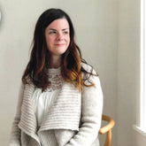 A woman with long dark hair and light highlights is sitting in a wooden chair, wearing a white lace top underneath the Knitbot Harborside Cardigan from Never Not Knitting. She is smiling softly and looking slightly to the side. The background is a minimalist, light-filled room.