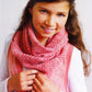 A young girl with long brown hair smiles while wearing the Ribbed Cowl from the Ella Rae Lace Merino Pattern collection by Knitting Fever / Euro Yarns. Her stylish ensemble includes a matching sleeveless top over a white long-sleeve shirt, adding a touch of elegance as she stands against a plain background.