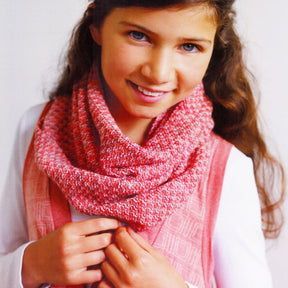 A young girl with long brown hair smiles warmly. She is wearing a stylish Ribbed Cowl – Ella Rae Lace Merino by Knitting Fever / Euro Yarns, draped over a white shirt and a pink, patterned vest. She holds part of the cowl with her hands while looking at the camera. The background is plain and light-colored.