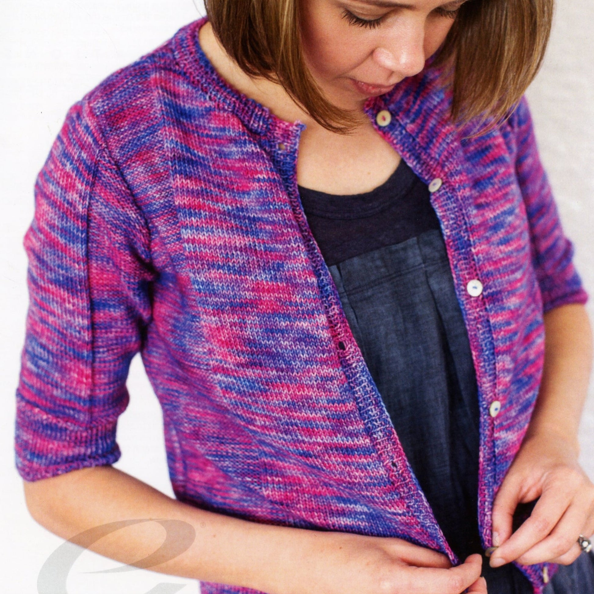 A person with shoulder-length brown hair is buttoning the charming Sara Cardigan by Knitting Fever / Euro Yarns, featuring a delightful Ella Rae Lace Merino pattern in shades of purple, pink, and blue. They are wearing a dark top underneath and are concentrating on fastening the buttons.