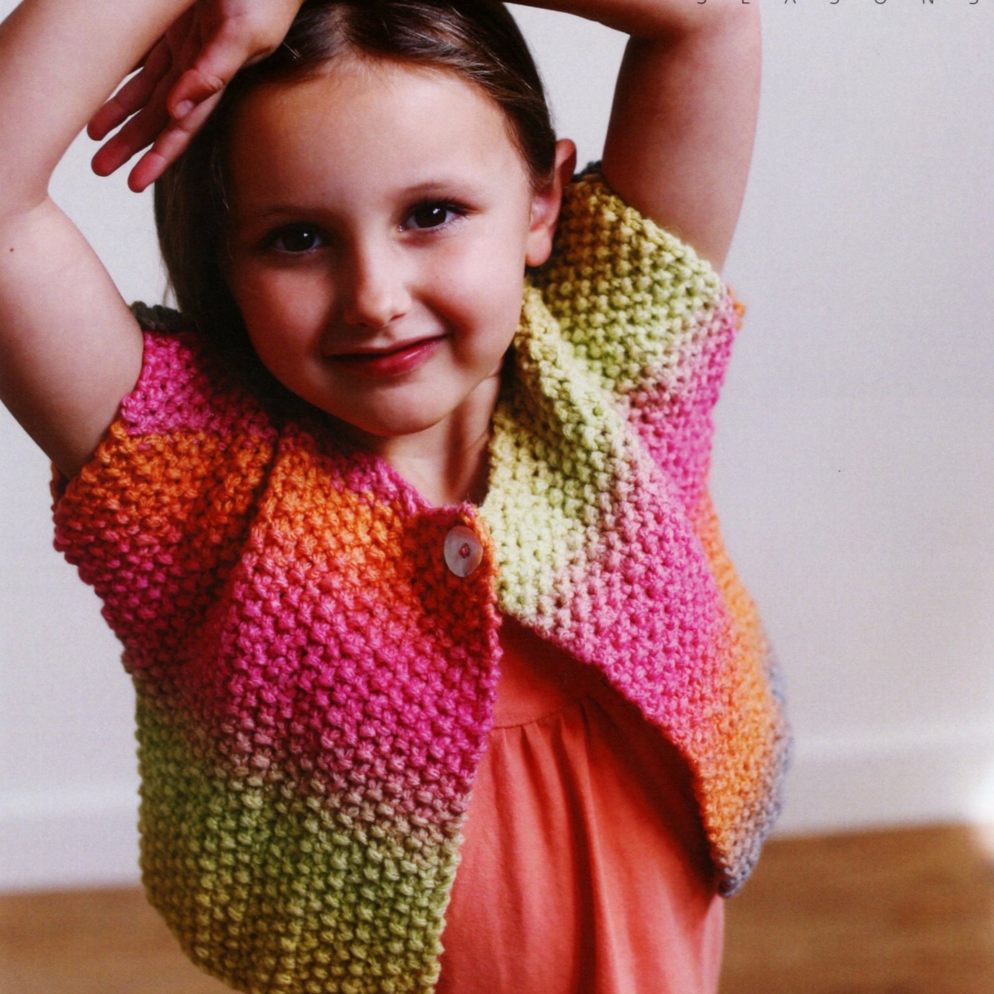 A young child with a short haircut, wearing a Moss Stitch Jacket from Ella Rae Seasons by Knitting Fever / Euro Yarns, smiles at the camera with arms raised. The jacket features a gradient pattern with shades of pink, yellow, orange, and green. The child is standing against a plain background.