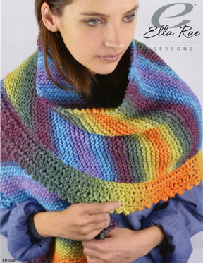 A person wearing a hand-knit, multicolored Ella Rae Seasons Amigo Shawl with hues of purple, green, blue, yellow, and pink. The garter stitch shawl is draped over their shoulders as they hold the edge while looking to the side. The words "Knitting Fever / Euro Yarns" are printed in the upper right corner.