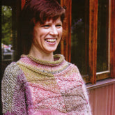 A person with short brown hair is smiling while wearing a Skeppsta Sweater from the Noro Revisited collection by Knitting Fever / Euro Yarns. The sweater features a vibrant, patchwork knit design. They stand in front of a wooden structure with windows, set against an artfully blurred background.