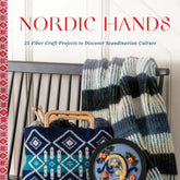 The book cover for "Nordic Hands" by Schiffer Publishing Co. showcases 25 fiber craft projects that beautifully capture Scandinavian culture. The cover features a knitted striped blanket casually draped over a chair, a blue patterned knitting project, and a round decorative tray adorned with floral designs, embodying the essence of Scandinavian life.