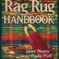 The cover of the "Rag Rug Handbook - 2nd Edition" by Janet Meany and Paula Pfaff from Great Northern Weaving showcases a vibrant woven rug background adorned with traditional patterns. At the center, wooden weaving tools and fabric strands vividly illustrate the art of weaving rag rugs. The title is prominently displayed at the top, while the authors' names are positioned at the bottom.