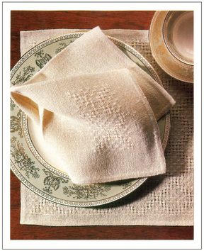 A neatly folded cream-colored napkin from the Handwoven Design Collection #11 by Long Thread Media lies on a decorative plate with a subtle floral pattern. The plate is set on a matching placemat atop a wooden table, accompanied by a ceramic bowl partially visible in the top corner.