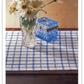 A clear vase filled with white daisies and a blue glass box rest on a wooden table adorned with the Handwoven Design Collection #11 by Long Thread Media, featuring heirloom table linens in a white and blue checkered pattern, set against a beige backdrop.