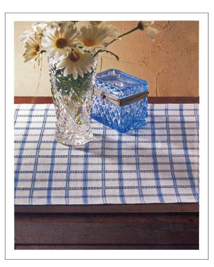 A clear vase filled with white daisies and a blue glass box rest on a wooden table adorned with the Handwoven Design Collection #11 by Long Thread Media, featuring heirloom table linens in a white and blue checkered pattern, set against a beige backdrop.