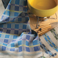 A vibrant yellow fondue pot on a stand is complemented by forks with wooden handles. The scene is enhanced with blue, green, and beige checkered cloths elegantly positioned next to it, all resting on a meticulously woven surface from the Handwoven Design Collection #18 by Long Thread Media.