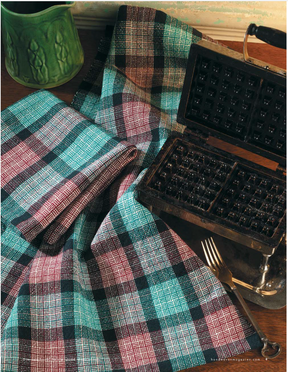 A handwoven design from Long Thread Media's "Collection #18, A Treasury of Towels" eBook is elegantly displayed beside an open vintage waffle iron. In the background, a green jug adds a splash of color, while a silver fork lies partially visible next to the iron on a wooden surface.