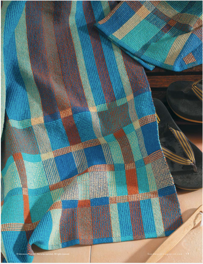 Displayed alongside a stylish pair of black sandals with gold detailing is the Handwoven Design Collection #18 from Long Thread Media. This printed version of 'A Treasury of Towels eBook' features a vibrant textile with a checkered pattern in blue, turquoise, brown, and orange hues. The fabric's rich texture and colors reflect light beautifully, creating an eye-catching visual effect.