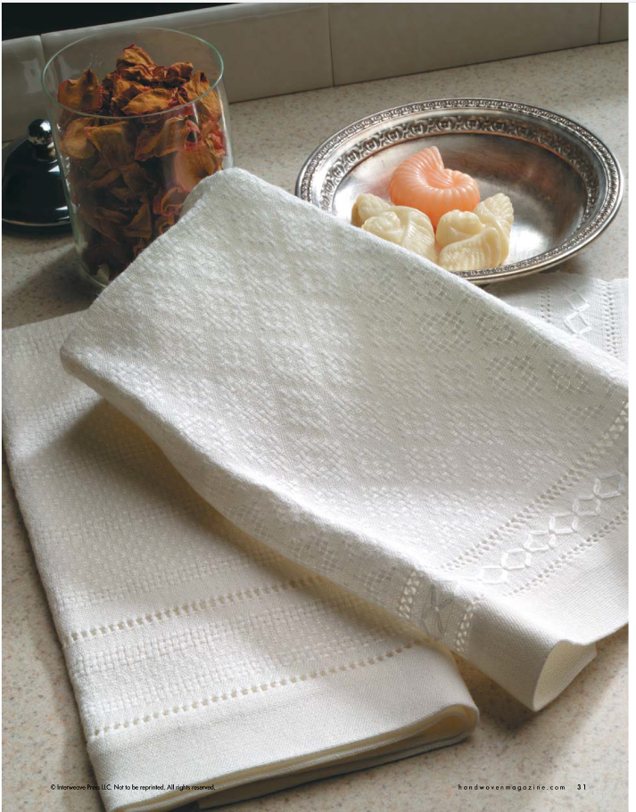 A neatly folded towel from the Handwoven Design Collection #18, highlighted in *A Treasury of Towels* by Long Thread Media, sits on a countertop beside a bowl of decorative soaps and a clear jar filled with dried rose petals. The soft lighting emphasizes the towel's intricate woven pattern.