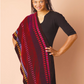 A person wearing a black outfit with a piece from Long Thread Media's Handwoven Design Collection #19, featuring vibrant striped shawls and scarves for all seasons, is draped over one shoulder, standing against a pale peach background while smiling and facing the camera.