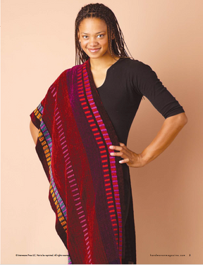 A person wearing a black outfit with a piece from Long Thread Media's Handwoven Design Collection #19, featuring vibrant striped shawls and scarves for all seasons, is draped over one shoulder, standing against a pale peach background while smiling and facing the camera.