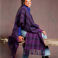 A person with a warm smile is sitting on a wooden beam, wrapped in a cozy purple and blue striped shawl from the Handwoven Design Collection #19 by Long Thread Media. They wear jeans and black boots, set against a soft, muted background.