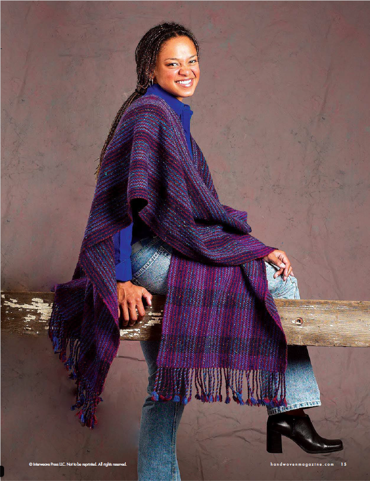 A person with a warm smile is sitting on a wooden beam, wrapped in a cozy purple and blue striped shawl from the Handwoven Design Collection #19 by Long Thread Media. They wear jeans and black boots, set against a soft, muted background.