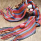 A woven basket from Long Thread Media's "Handwoven Design Collection #19, Scarves and Shawls for All Seasons eBook (Printed version)" contains four colorful eggs and a striped scarf with fringe in shades of orange, red, purple, and gray. The basket and scarf are placed on a wooden surface, with one egg delicately resting on the scarf—an inspiring example of creative weave projects.