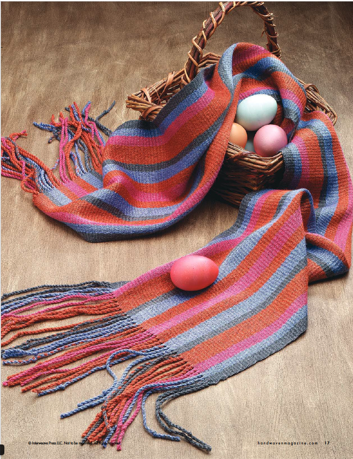 A woven basket from Long Thread Media's "Handwoven Design Collection #19, Scarves and Shawls for All Seasons eBook (Printed version)" contains four colorful eggs and a striped scarf with fringe in shades of orange, red, purple, and gray. The basket and scarf are placed on a wooden surface, with one egg delicately resting on the scarf—an inspiring example of creative weave projects.