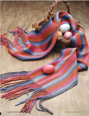 A woven basket from Long Thread Media's "Handwoven Design Collection #19, Scarves and Shawls for All Seasons eBook (Printed version)" contains four colorful eggs and a striped scarf with fringe in shades of orange, red, purple, and gray. The basket and scarf are placed on a wooden surface, with one egg delicately resting on the scarf—an inspiring example of creative weave projects.