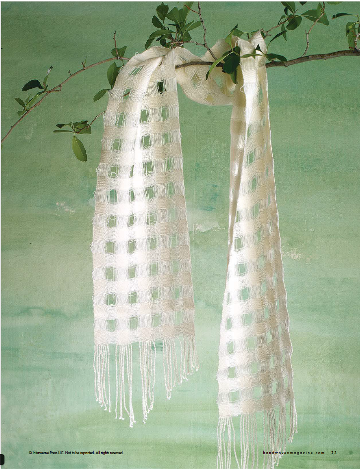 A delicate, handwoven scarf from Halcyon Yarn's Handwoven Design Collection #19 by Long Thread Media drapes gracefully over a thin branch against a soothing green backdrop. The white scarf features a checkered pattern and silky texture, enhanced by fringe details at the ends, exemplifying the creative craftsmanship found in woven projects for all seasons.