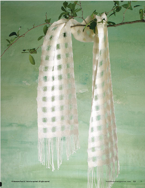 A delicate, handwoven scarf from Halcyon Yarn's Handwoven Design Collection #19 by Long Thread Media drapes gracefully over a thin branch against a soothing green backdrop. The white scarf features a checkered pattern and silky texture, enhanced by fringe details at the ends, exemplifying the creative craftsmanship found in woven projects for all seasons.